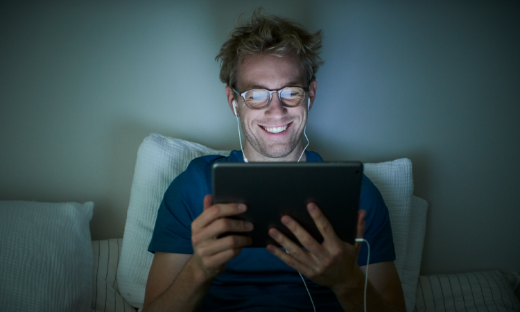 Jeune homme allongé dans son lit et exposé à de la lumière émise par l'écran de sa tablette tactile, en intérieur.